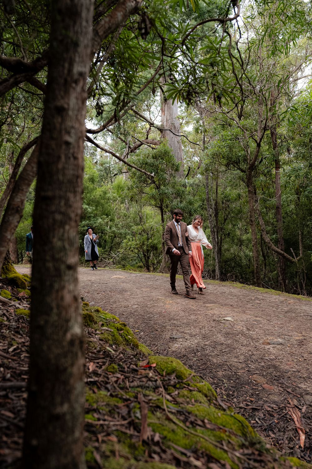 A couple walking along a mossy forest trail with tall eucalyptus trees creating a natural canopy.