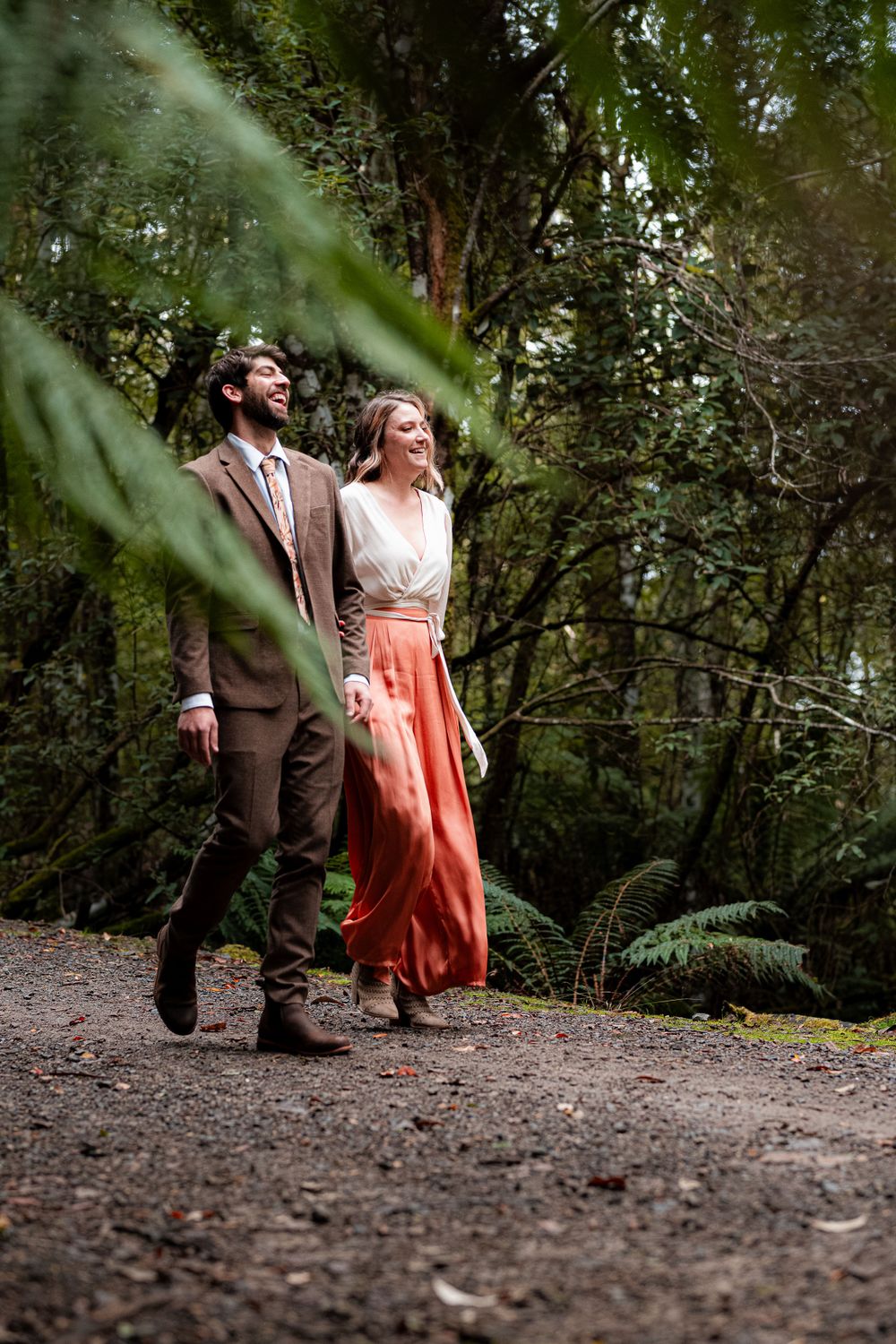 A couple walking together on a forest path wearing formal attire amidst green foliage.