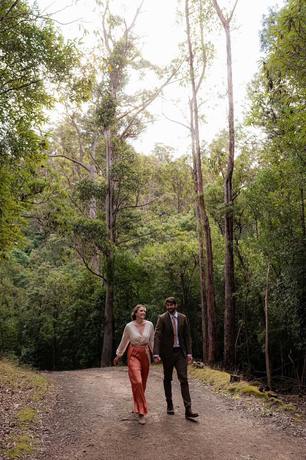 A couple walks together on a forest trail surrounded by tall eucalyptus trees on a sunny afternoon.