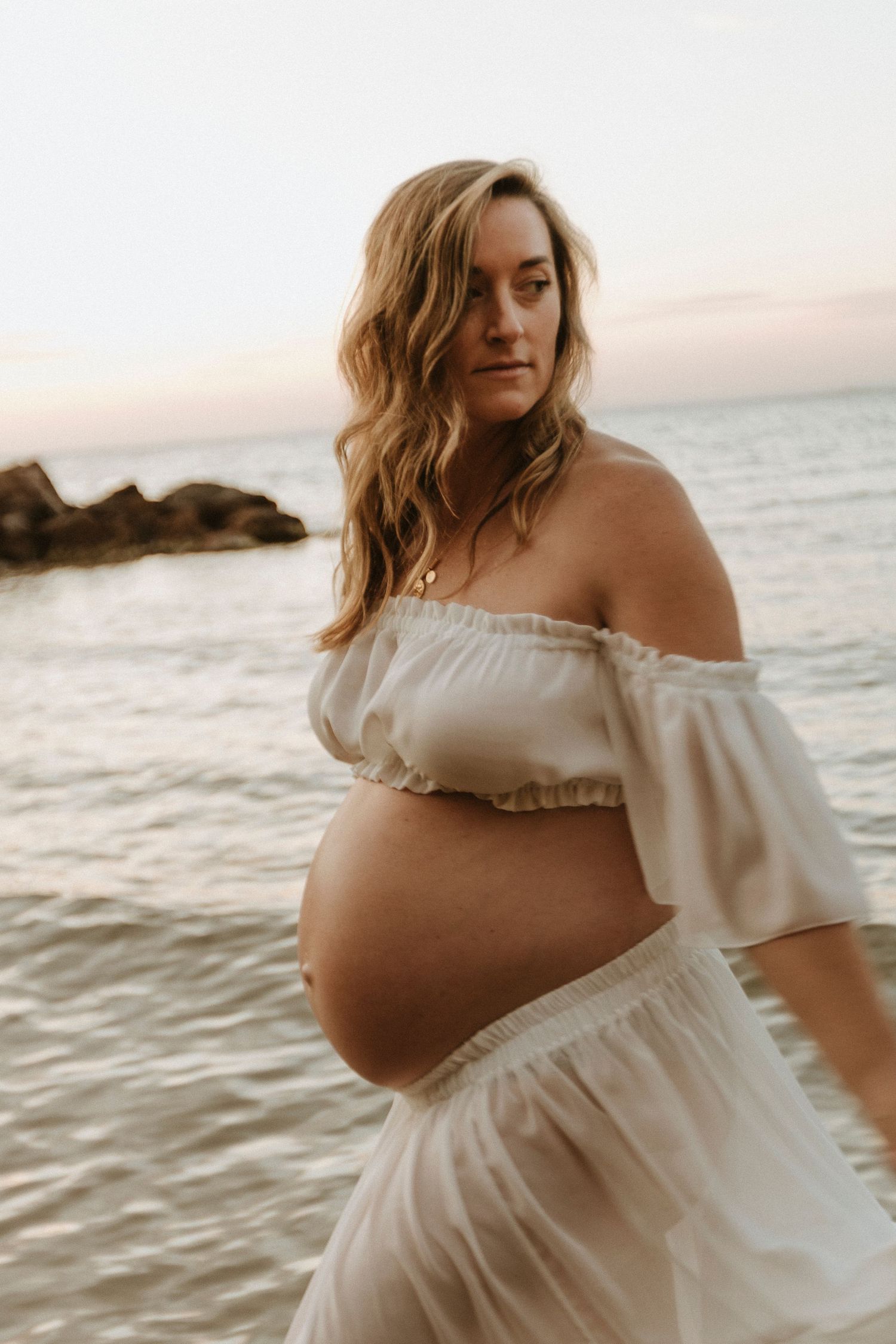 Pregnant woman in white off-shoulder dress poses at beach during sunset in maternity photoshoot series.