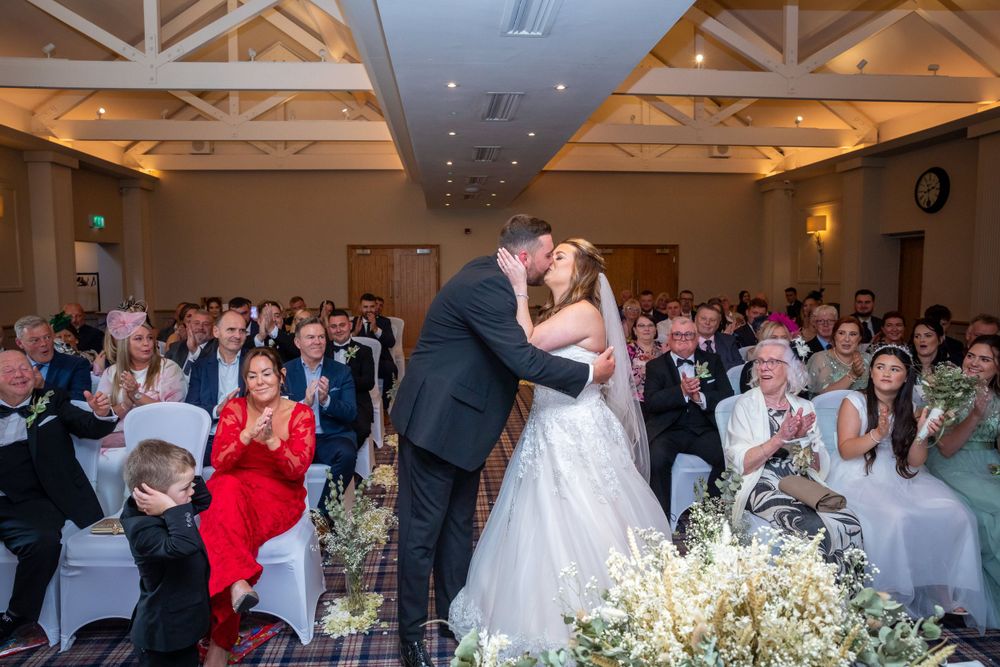Bride and groom share first kiss at wedding ceremony while guests applaud in elegant venue.