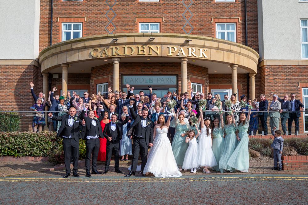 Wedding party and guests pose outside Carden Park Hotel for large group celebration photo.