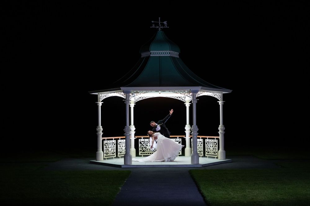 Bride and groom dance romantically inside illuminated white gazebo at night.