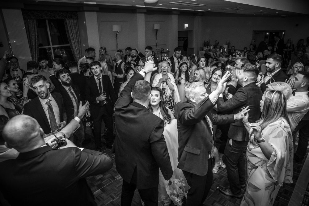 Bride and groom celebrate with guests on busy wedding reception dance floor in black and white.