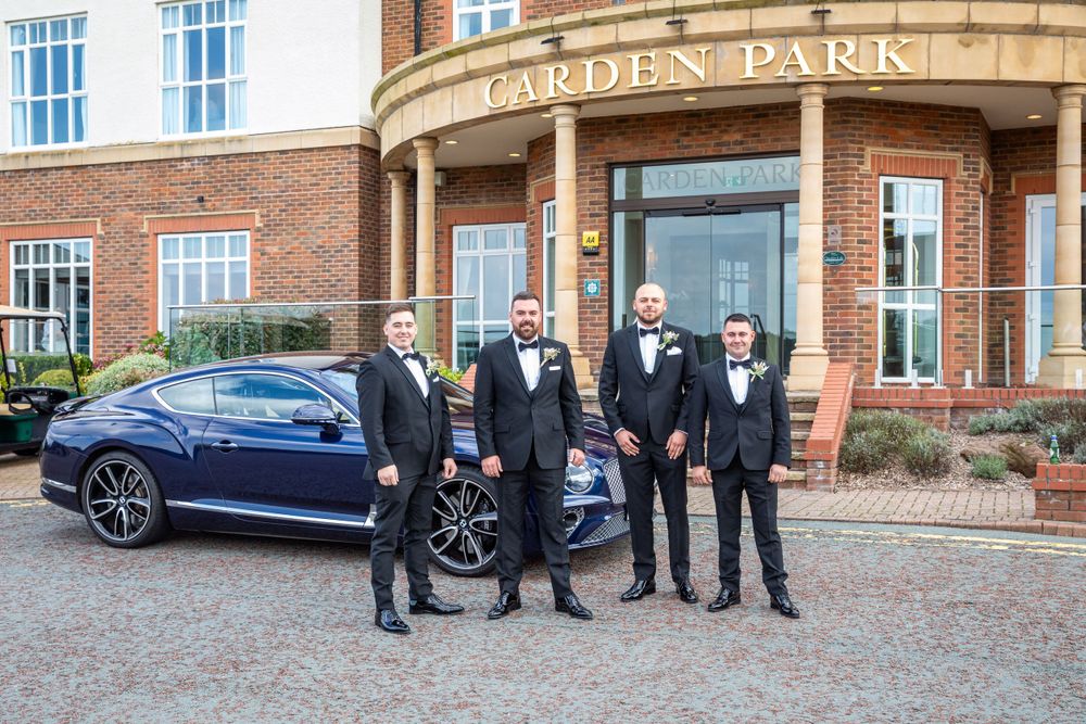 Groom and groomsmen in tuxedos posing beside a blue Bentley outside Garden Park Hotel wedding venue.
