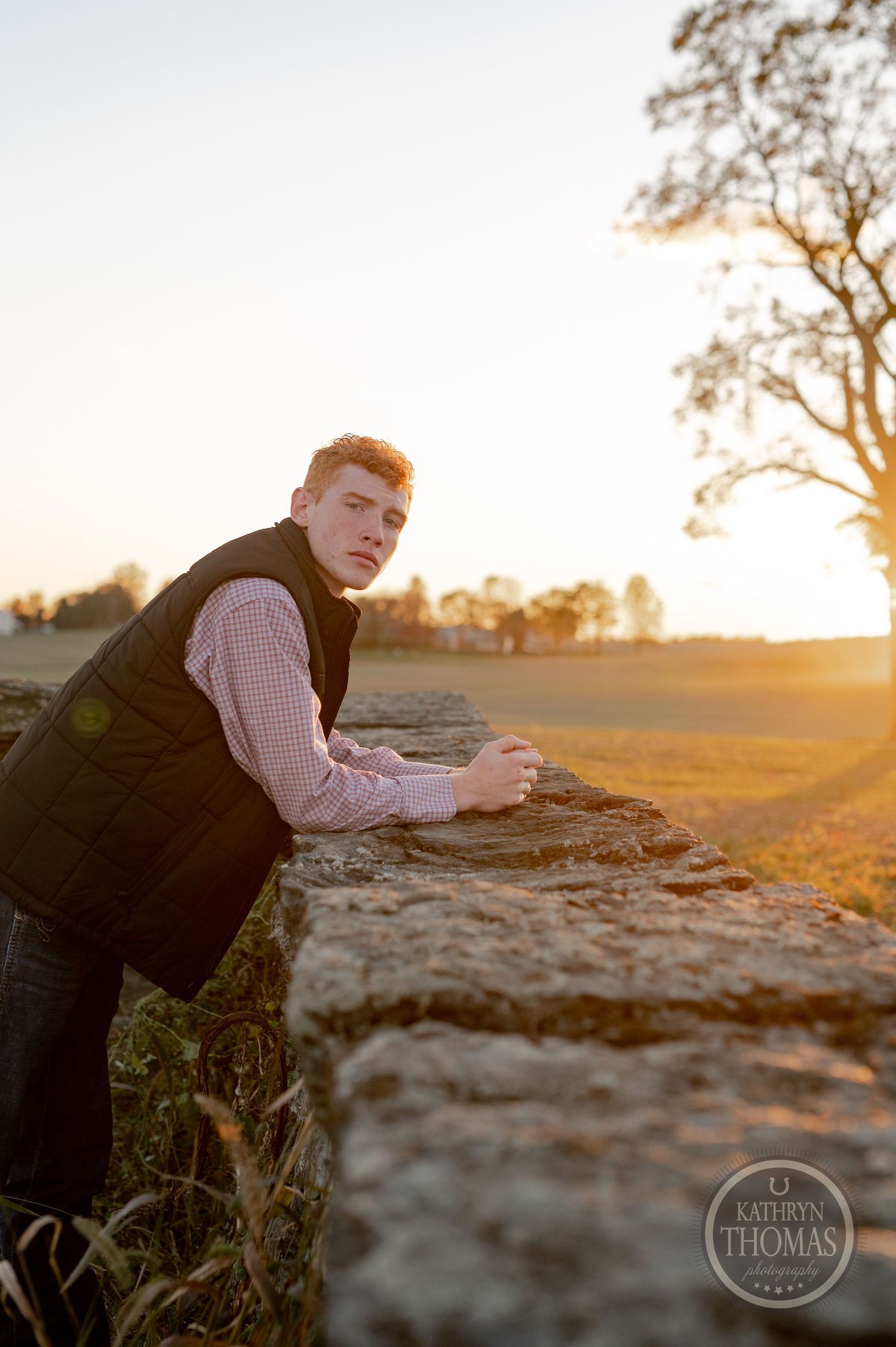 Person in vest and button-down shirt leaning on stone wall during golden hour.