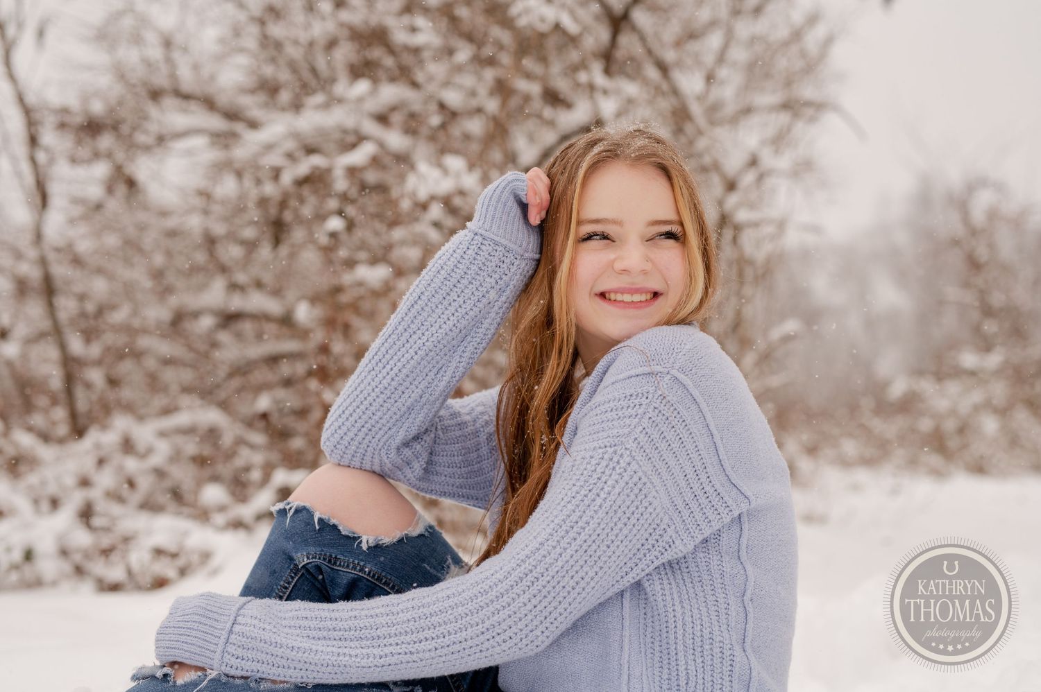 A person in winter clothing embraces their knees while sitting in snow surrounded by trees.