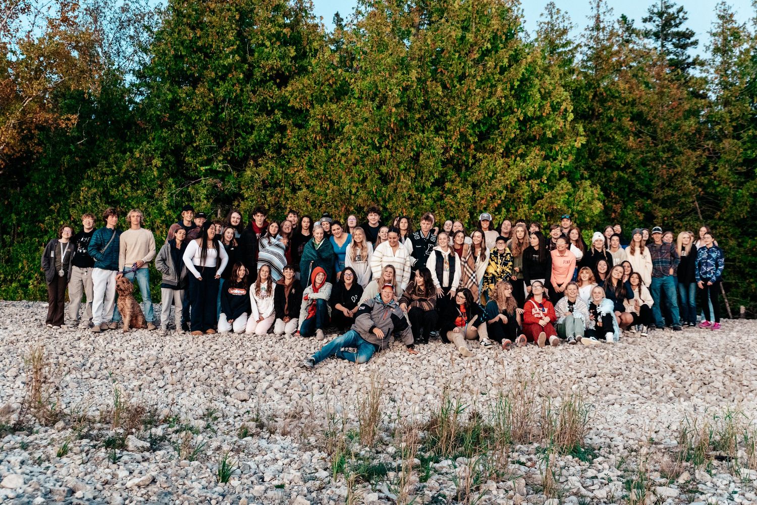 Large group photo of people gathered on rocky beach with evergreen trees in background.