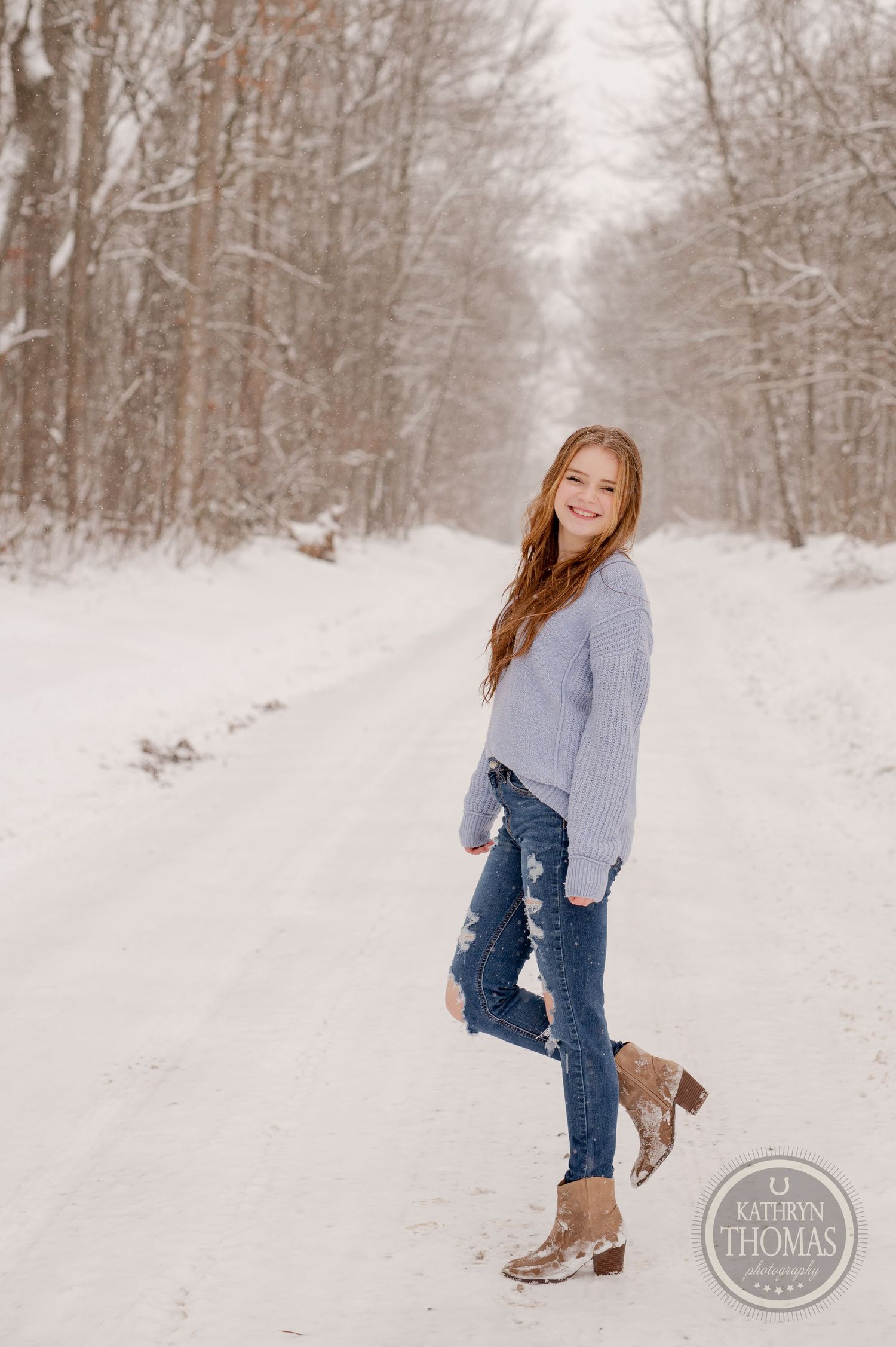 A person in a gray sweater and ripped jeans stands on a snowy forest path wearing brown boots.