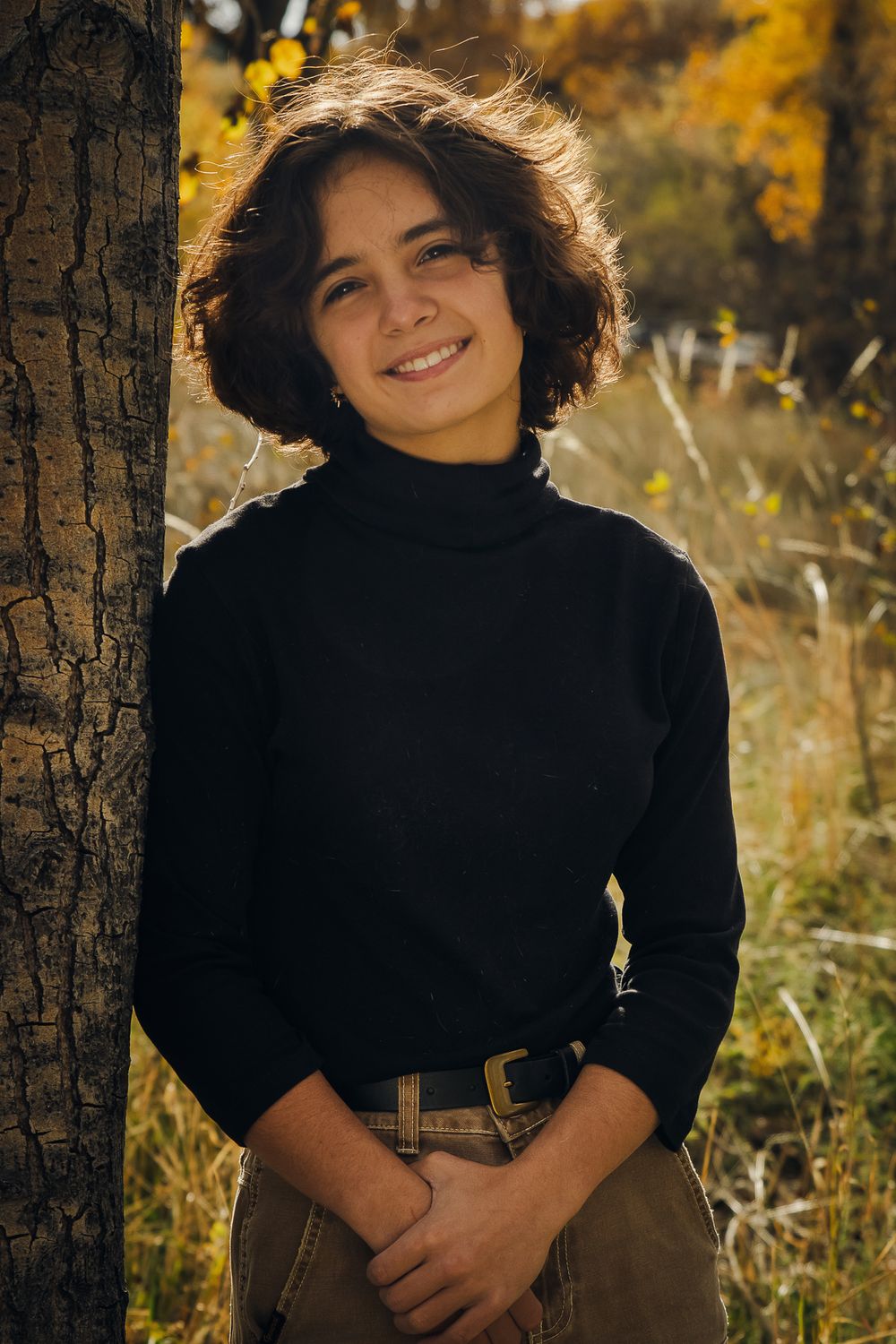 A person in a black sweater poses outdoors against autumn foliage and tree trunks in warm golden light.