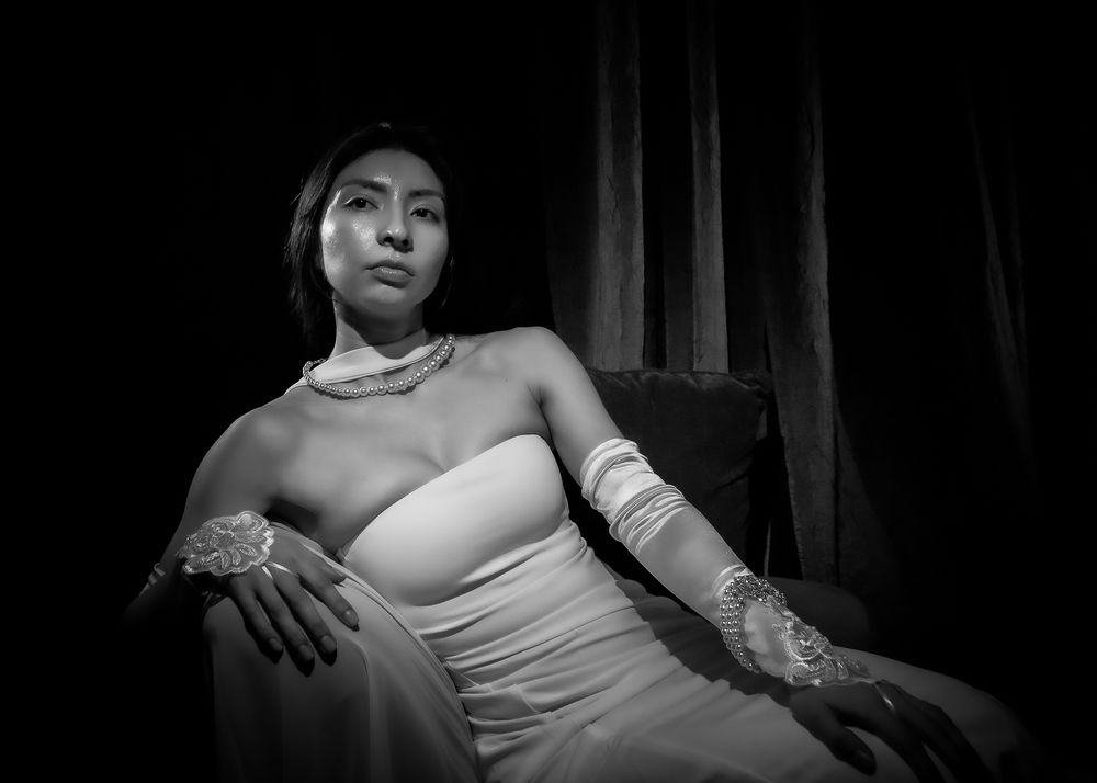 Black and white portrait of a young woman in elegant white gown and lace gloves, seated gracefully in dramatic lighting.