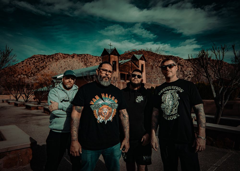 Four tattooed rock band members pose outdoors with red mountains and a historic building backdrop.