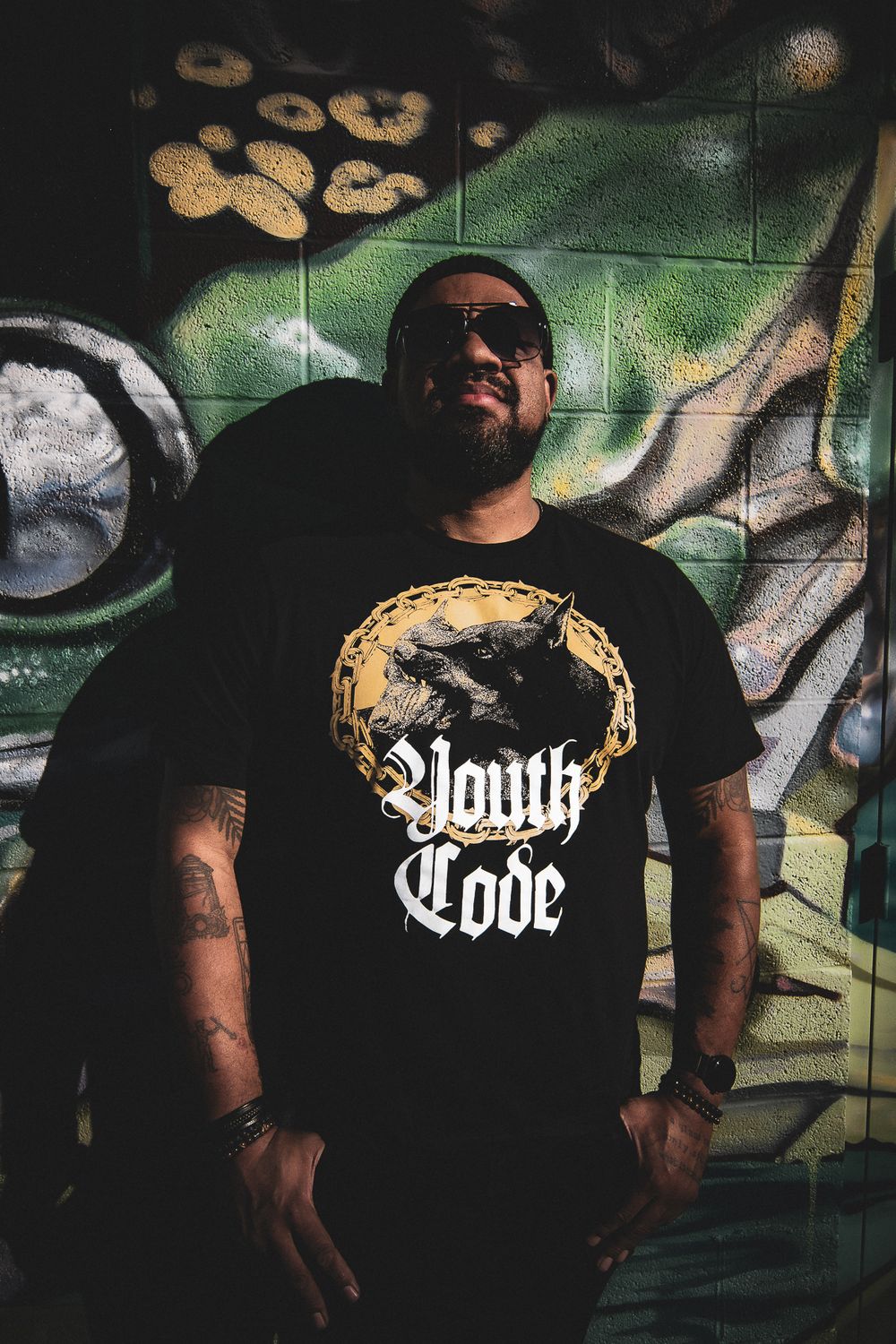 A person wearing sunglasses and a black Youth Code t-shirt stands against a graffiti wall with urban street art.