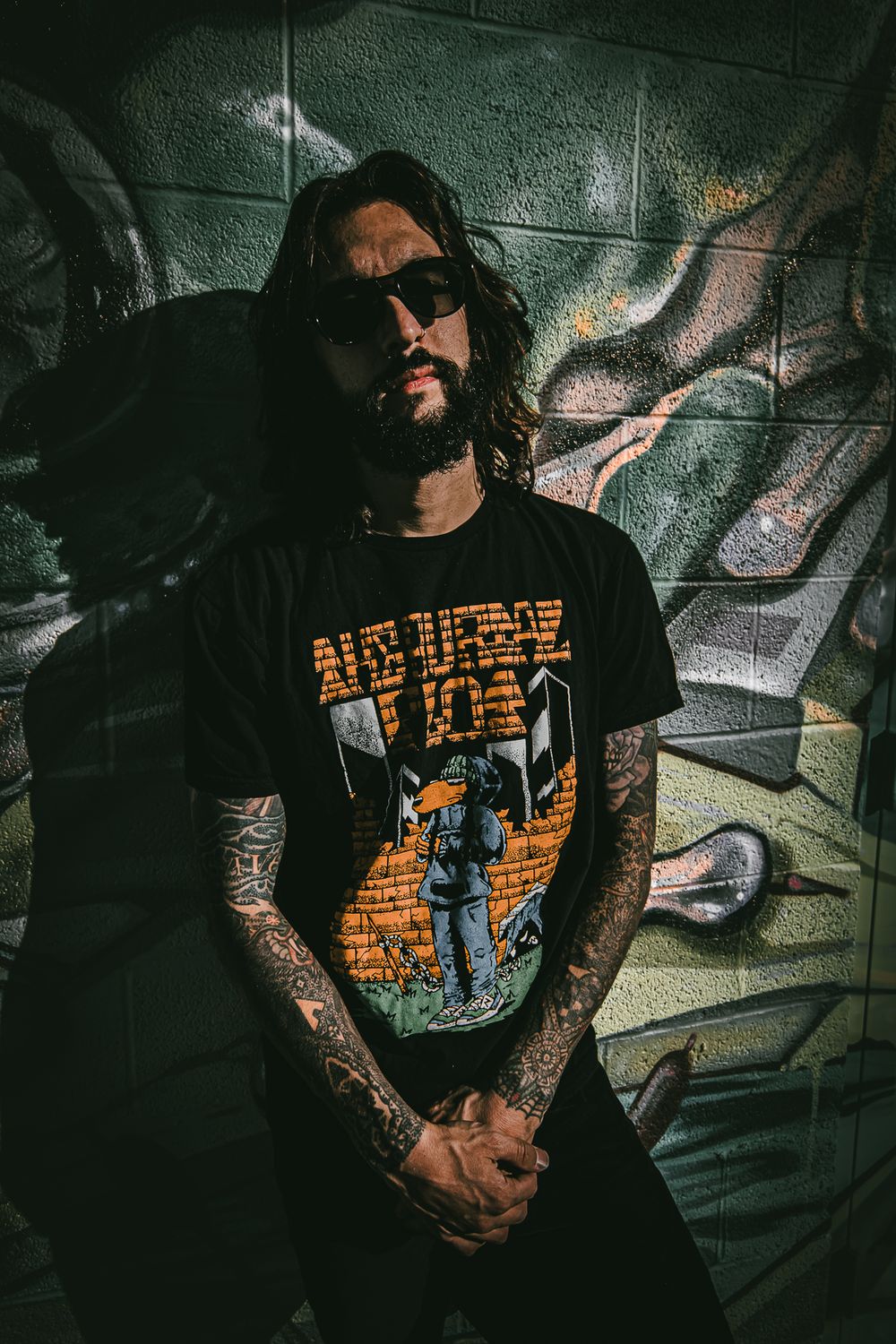 Tattooed figure in black t-shirt with orange design poses against graffiti wall in moody lighting.