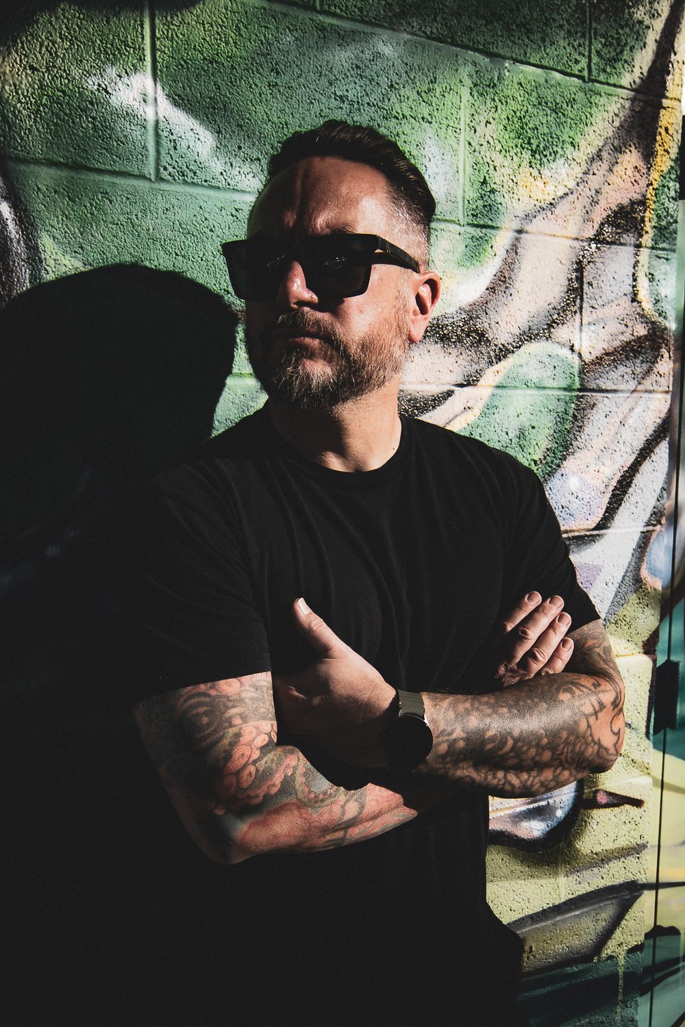Someone in sunglasses and black t-shirt with tattoos poses with crossed arms against graffiti wall in moody lighting.