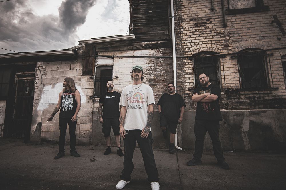 Band members posing against industrial brick wall backdrop in moody black and white.