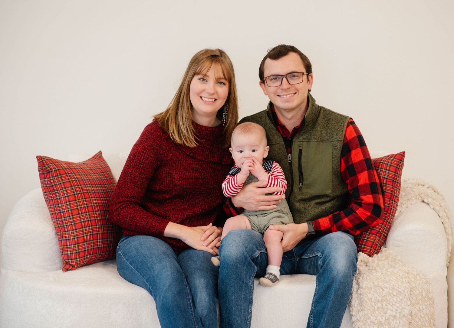 A happy family of three poses on a white couch, with parents holding their baby, wearing festive red and plaid outfits.