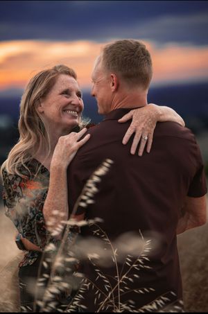 Couple embracing at sunset in a golden field, smiling at each other with a colorful sky backdrop.