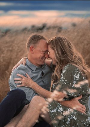 A couple shares a romantic moment sitting in a golden field at sunset, smiling and embracing each other closely.