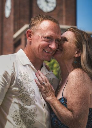 Couple sharing a romantic moment outdoors near a brick building with a clock tower, man in white embroidered shirt smiling.