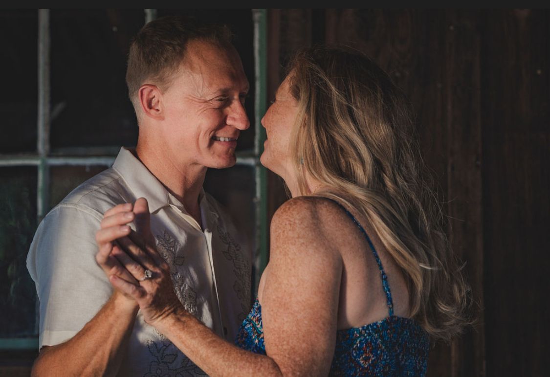 A smiling couple dances together in a warmly lit rustic setting, sharing a joyful and intimate moment.