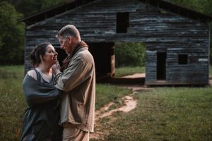 Couple embracing romantically in front of a weathered rustic barn in a field.