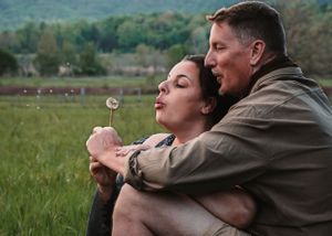 Couple sitting in a green field, woman blowing a dandelion while man embraces her from behind.