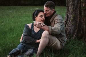 Couple sitting together against a tree in a grassy field, sharing an intimate embrace.