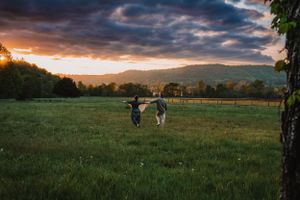 Couple running hand-in-hand through a green meadow at sunset with dramatic cloudy sky.