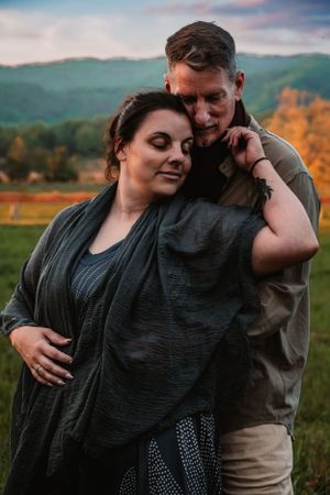 Couple embracing in a field with stunning mountain and autumn foliage backdrop at sunset.