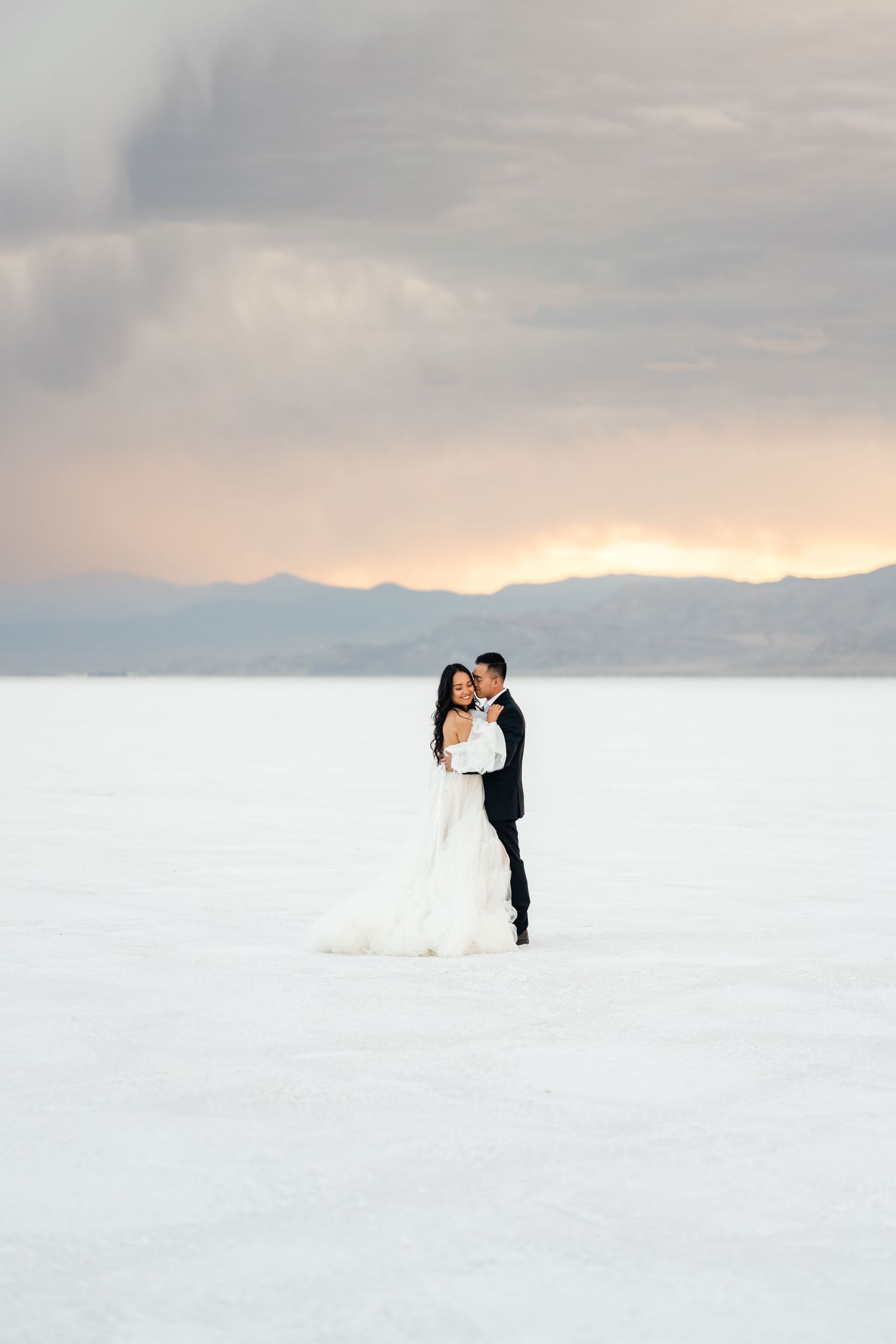 A couple in wedding attire standing on white salt flats at sunset.