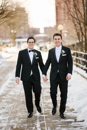 Two grooms in black tuxedos hold hands walking on a snowy urban path, smiling on their winter wedding day.