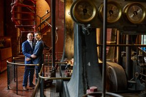 Two grooms embrace on a balcony inside a historic industrial venue with vintage machinery and spiral staircase.