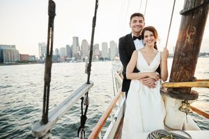 Bride and groom embrace on a sailboat with Boston city skyline and harbor in the background on their wedding day.