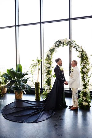Same-sex couple exchanging vows under a white floral arch, one in black gown with train, other in white suit.