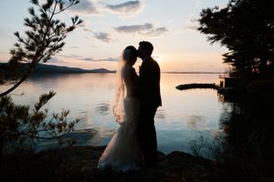 Silhouette of bride and groom sharing a kiss at sunset by a calm lake with mountains in the background.