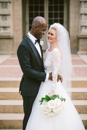 Interracial couple in wedding attire, groom in tuxedo and bride in lace gown with veil, holding white roses bouquet.