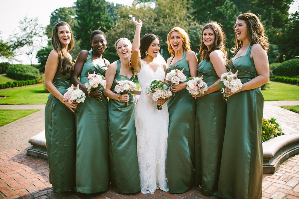 A bride throwing her hand up and laughing standing in a rose garden with her bridesmaids in green all laughing along with her.