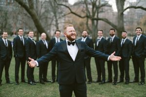 Groom in black tuxedo poses with arms wide open in front of groomsmen in a park setting.