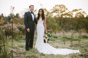 Bride in white gown holding bouquet stands with groom in black tuxedo in a natural outdoor field setting.