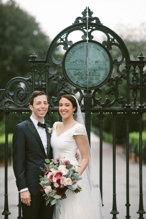 Bride and groom smiling in front of ornate Boston Public Garden iron gates, bride holding lush pink and burgundy bouquet.