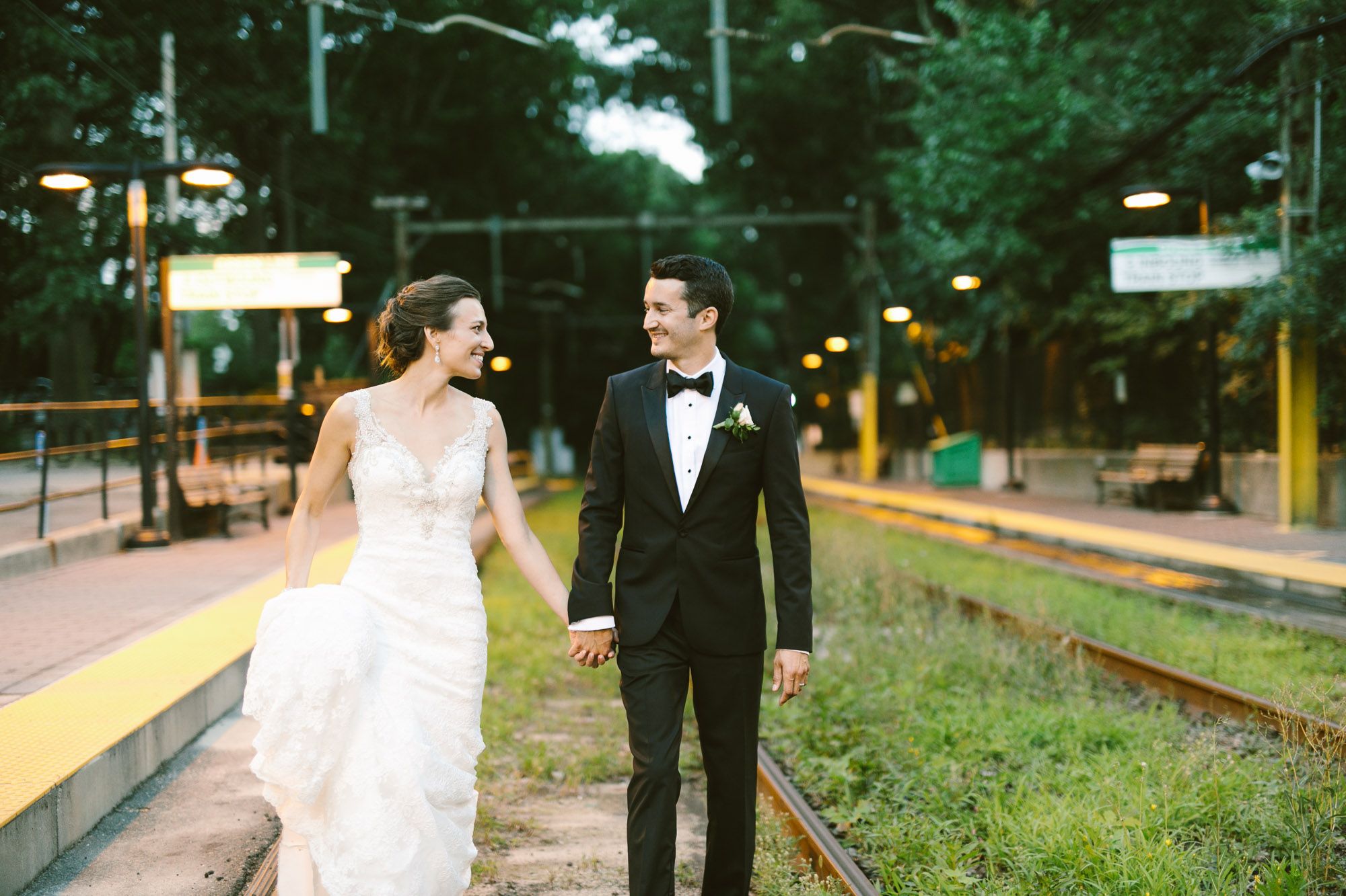Bride and groom hold hands smiling at each other near railroad tracks at golden hour in elegant wedding attire.