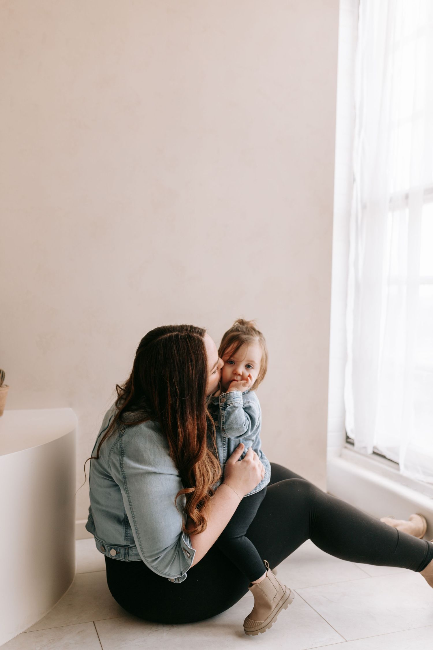 A young mother in a denim jacket sits on a floor by a window, tenderly kissing her smiling toddler daughter.