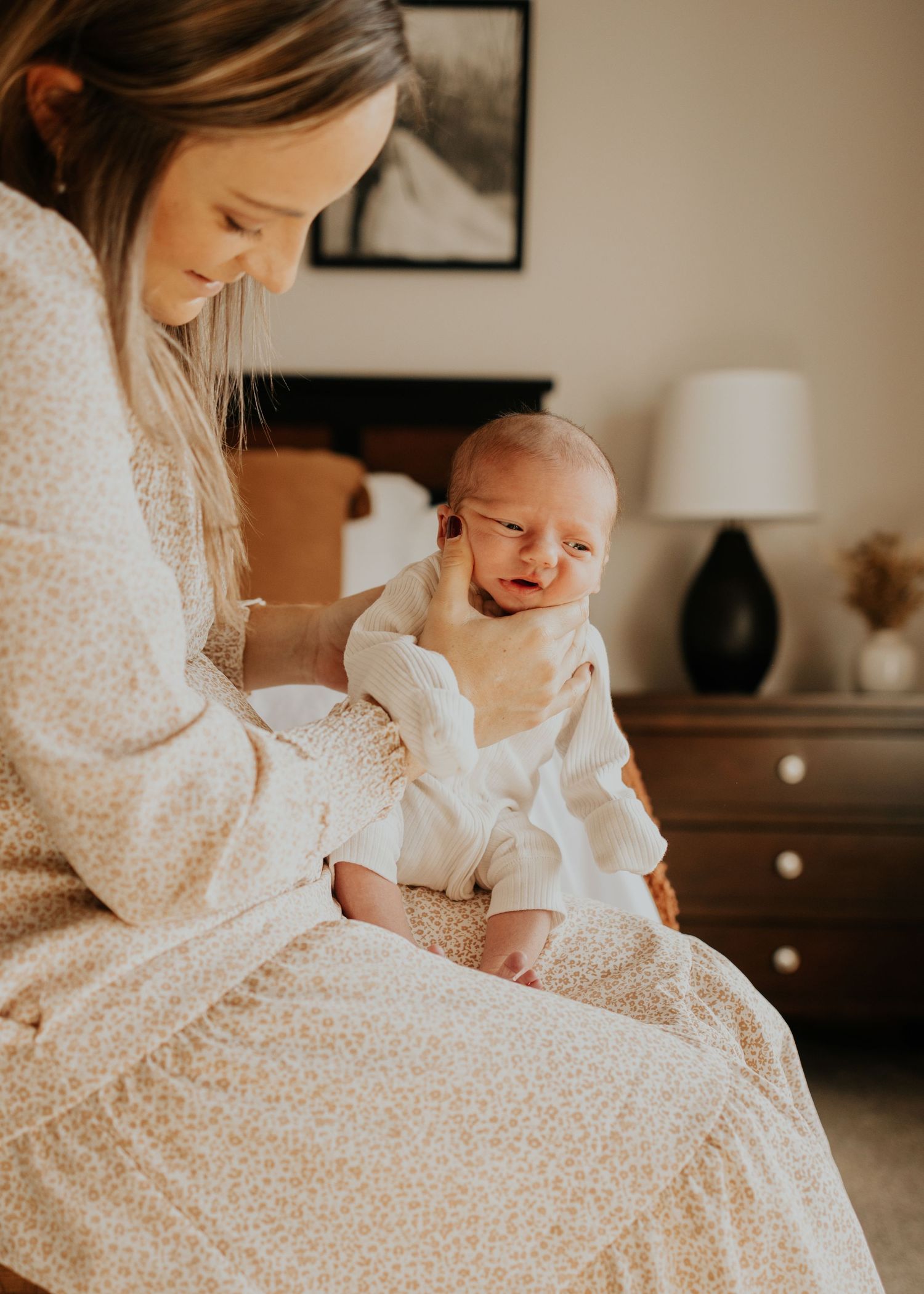 A crying baby in white clothing sits on a bed with a caregiver in a warm-lit bedroom interior.