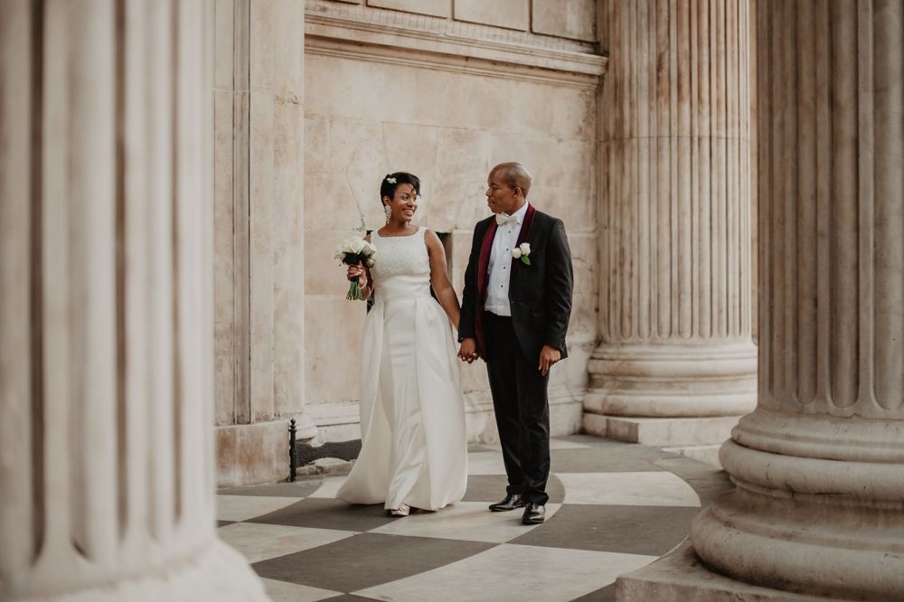Bride and groom hold hands walking between towering neoclassical columns. Bride in white strapless gown, groom in black suit