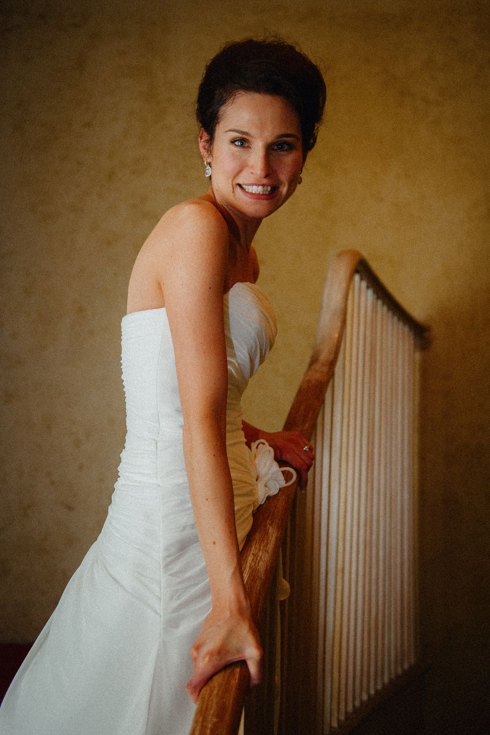 Smiling bride in strapless white gown descends wooden staircase. Portrait shows elegant updo hairstyle and pearl earrings indoors