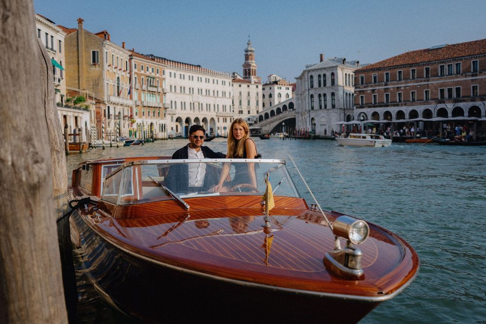 Couple aboard classic wooden motorboat on Venice's Grand Canal with Rialto Bridge and historic Venetian palazzos under blue sky
