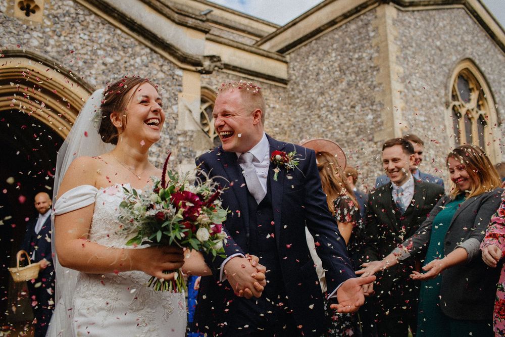 Laughing newlyweds exit stone church as wedding guests throw confetti. Bride in white dress with veil, groom in navy suit outside