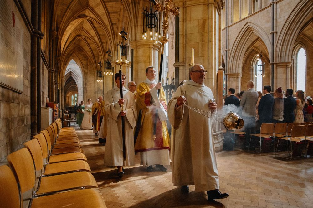 Wedding procession in Gothic cathedral with clergy in ceremonial robes. Orange chairs line nave under soaring stone arches