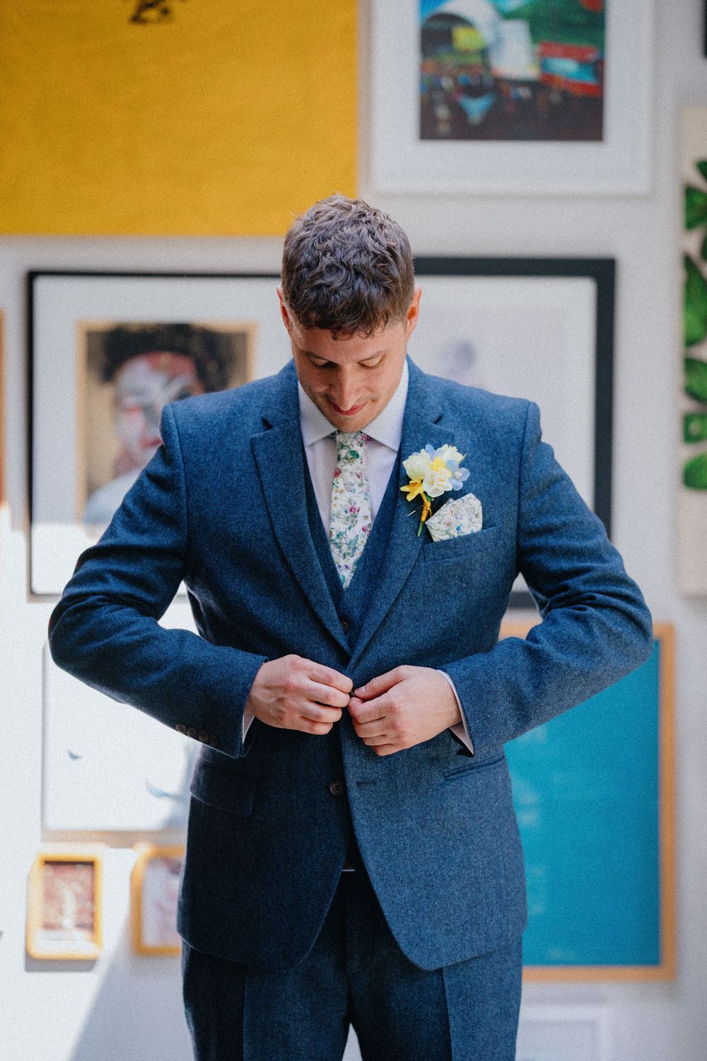 Groom in blue three-piece suit adjusts jacket buttons in modern interior with colorful artwork. White flower boutonniere visible